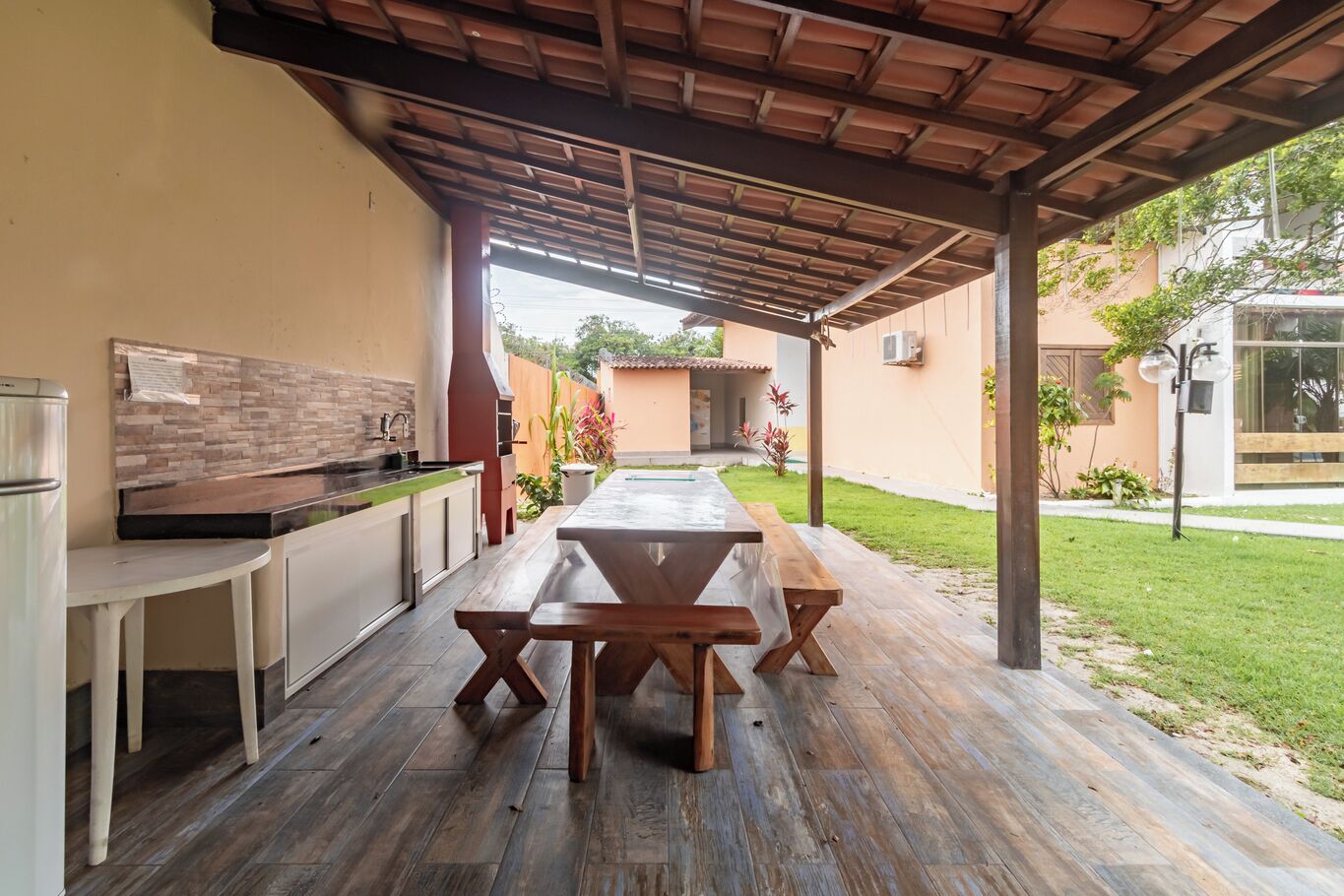 Um pátio externo com mesa e bancos de madeira, cozinha planejada com painel de pedra e área coberta com telhado em cerâmica.