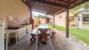 Um pátio externo com mesa e bancos de madeira, cozinha planejada com painel de pedra e área coberta com telhado em cerâmica.