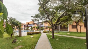 Uma trilha leva a uma área de piscina com espreguiçadeiras e guarda-sóis, tudo cercado por vegetação e palmeiras.