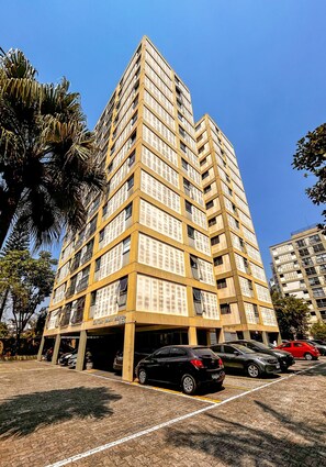 Um edifício alto, com vários andares e fachada uniforme, com varandas e área de estacionamento em frente.