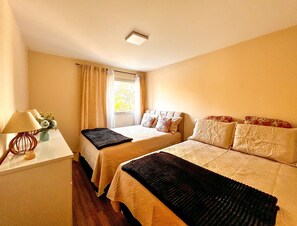 Quarto de hotel com duas camas, uma mesa com luminária e uma janela com cortinas.