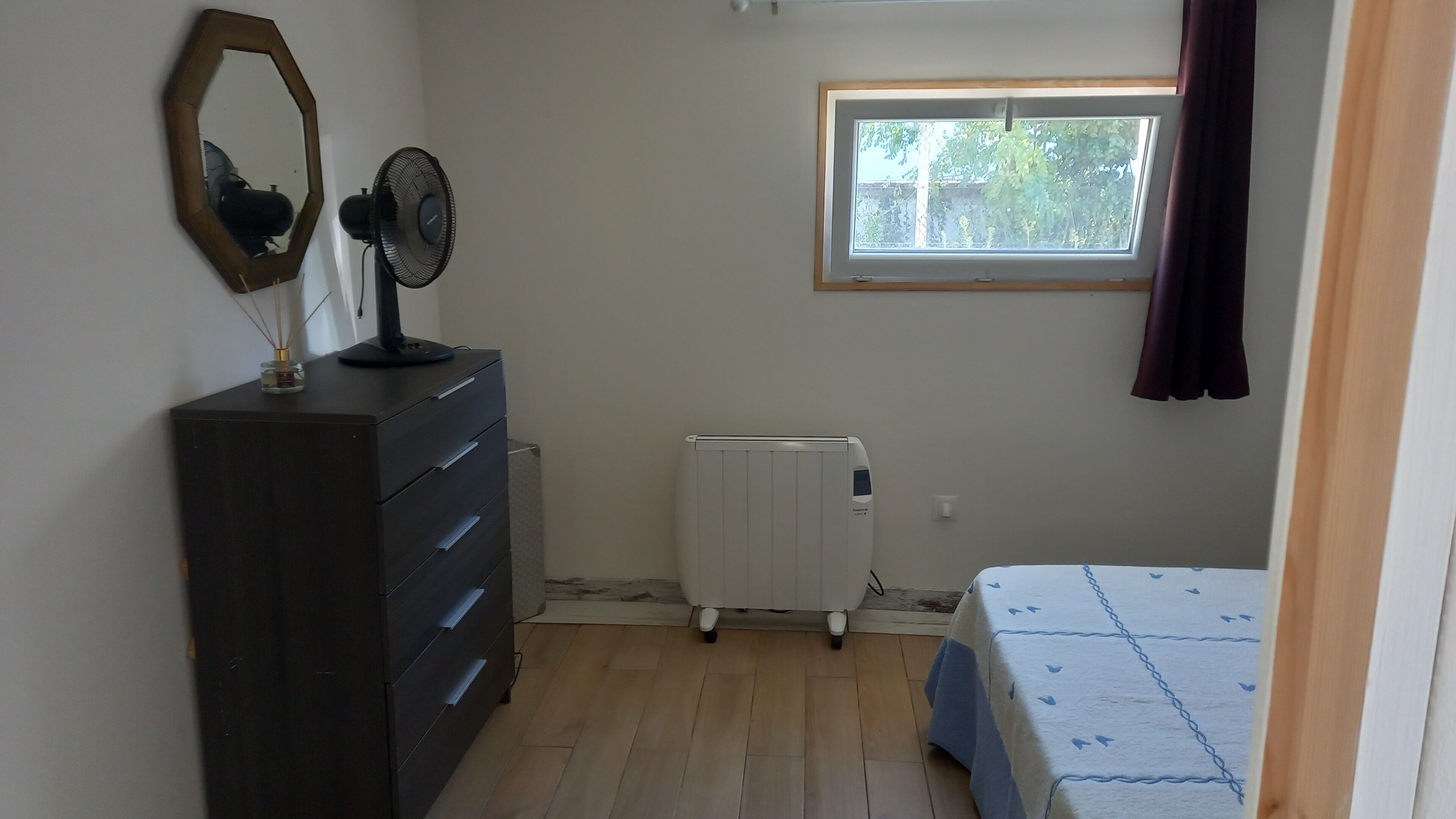 A bedroom with a dresser, a standing fan, a radiator, a window with a view of greenery, and a bed with a blue and white patterned bedspread.