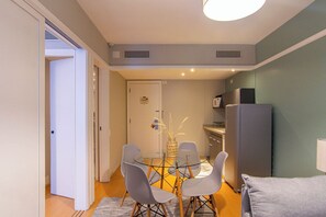 Ein kompakter Wohnbereich mit Essplatz, ausgestattet mit einem Glastisch und vier Stühlen, einer Küchenzeile inklusive Kühlschrank und einer Tür zu einem weiteren Raum.