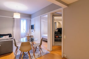 Ein modernes Wohnzimmer mit einem Glastisch, einer Couch und einem Fernseher. Ein Flur führt zu einem Schlafzimmer mit einem Bett und einem Kleiderschrank.