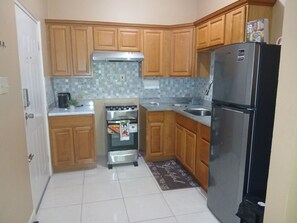 A compact kitchen with wooden cabinets, a stainless steel oven, a refrigerator, and a tiled backsplash. - OHC The Oasis at Hamlet C (Kingston)