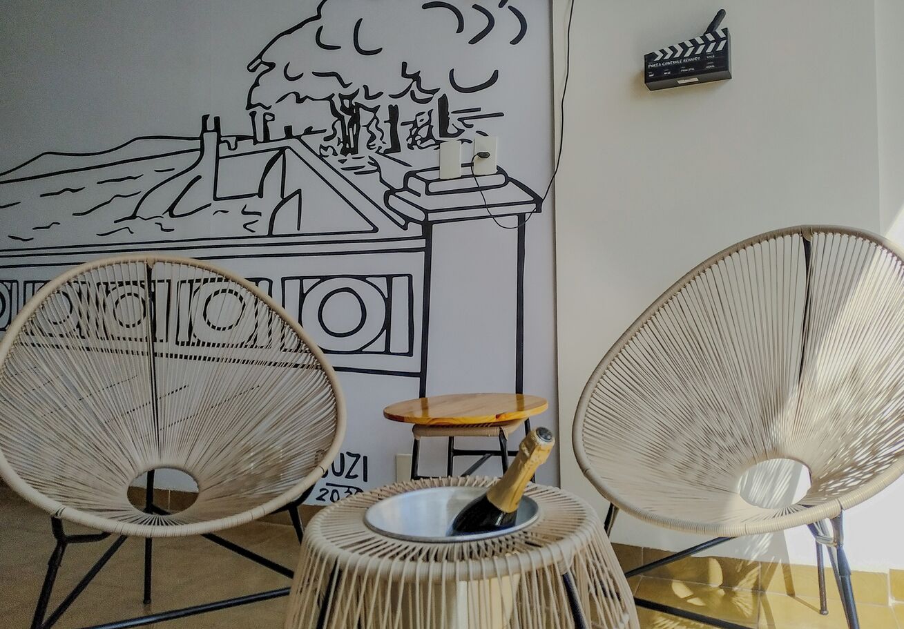Duas cadeiras de vime com uma pequena mesa de madeira no centro, uma garrafa de champanhe e um mural em preto e branco na parede.