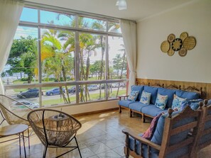 A room with a large window, a blue sofa, and a wicker chair. - Apto completo no Embaré: Beira Mar Ar Garagem (Santos)