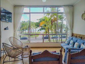 A room with a large window, a blue sofa, wicker chairs, and a wooden bed. - Apto completo no Embaré: Beira Mar Ar Garagem (Santos)