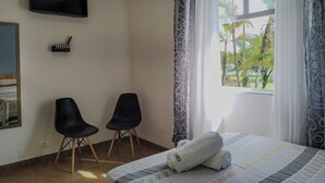 Ein Hotelzimmer mit einem Bett, zwei Stühlen, einem Flachbildfernseher und einem Fenster mit Blick auf Palmen.