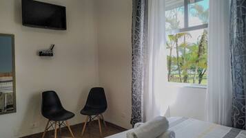 Quarto de hotel com cama, duas cadeiras, televisão de tela plana e janela com vista para palmeiras.
