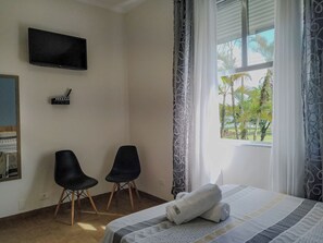 A hotel room with a bed, two chairs, a flat-screen TV, and a window with a view of palm trees. - Apto completo no Embaré: Beira Mar Ar Garagem (Santos)