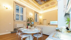 A dining area with a round table set for six, surrounded by clear chairs. There is a decorative plant centerpiece, a vase with flowers, and a television mounted on the wall. - Bella Benci Frescoed Apartment Central Florence near Uffizi and Ponte Vecchio (Firenze)