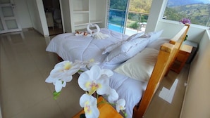 A hotel room with a large bed, a wooden nightstand, and a view of a mountainous landscape through a large window. - Bella vista Hotel  (Choachí)