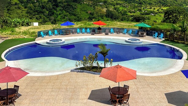A pool area with lounge chairs and umbrellas, surrounded by lush green hills and a clear blue sky. - Bella vista Hotel  (Choachí)