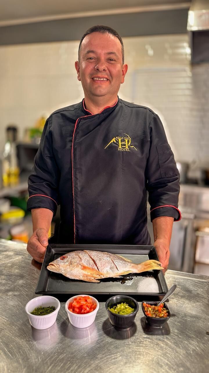 A chef in a black jacket with a logo, holding a tray with a fish and four small bowls of condiments.