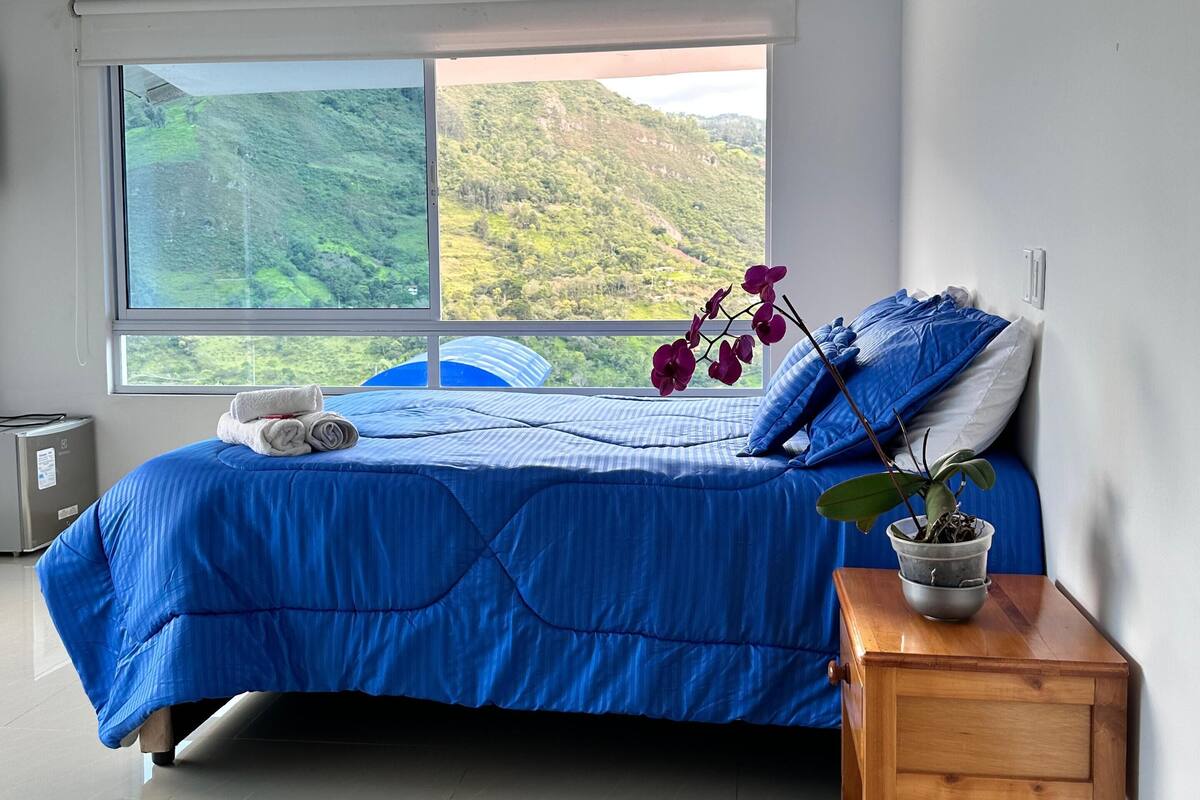 Een slaapkamer met een groot raam, een bed met blauw beddengoed, een houten nachtkastje en een potplant.