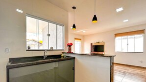 A modern kitchen with a large island, a built-in sink, pendant lighting, and a view through a large window. - Casa c/ Piscina 70m da Praia do Coral | Unamar esp (Cabo Frio)