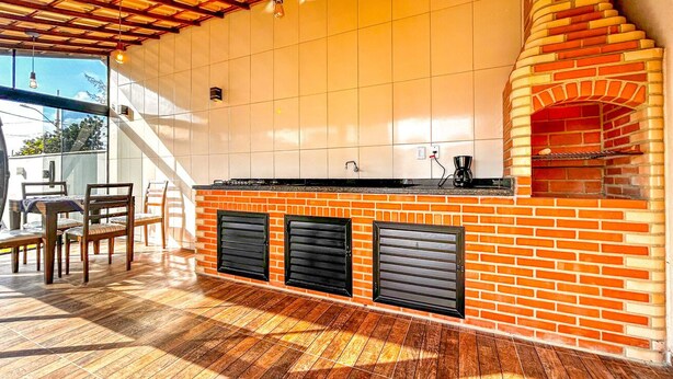 An outdoor kitchen area with a brick oven, wooden flooring, and a dining set. - Casa c/ Piscina 70m da Praia do Coral | Unamar esp (Cabo Frio)