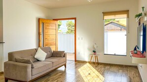 A living room with a beige sofa, wooden flooring, a small side table with a potted plant, and a window with a view of a house. - Casa c/ Piscina 70m da Praia do Coral | Unamar esp (Cabo Frio)