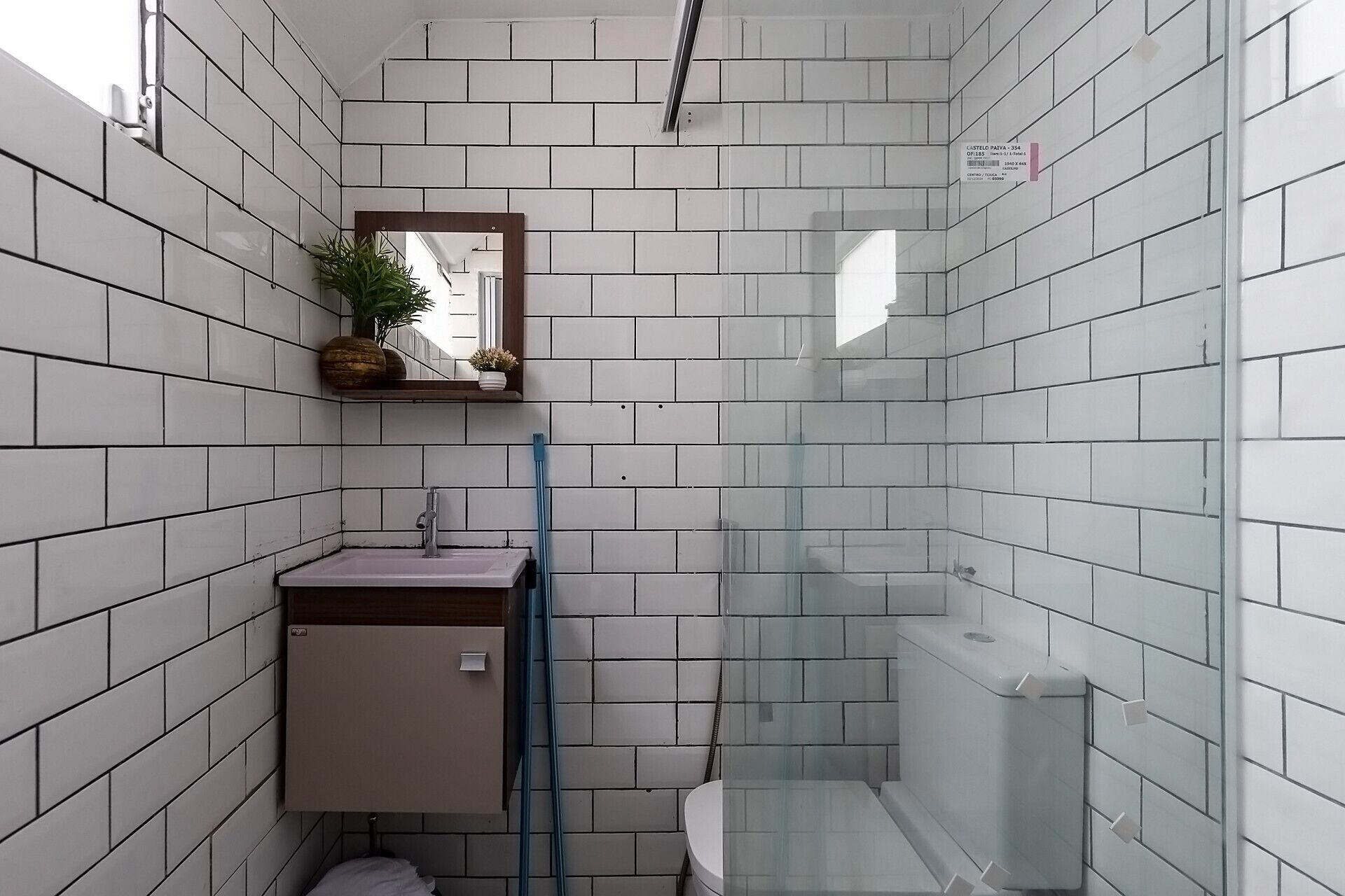Une petite salle de bain aux murs recouverts de carreaux blancs, avec un petit lavabo, un miroir et une cabine de douche en verre.