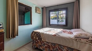 A bedroom with a bed, a window with curtains, a radiator, and a small table with a radio.