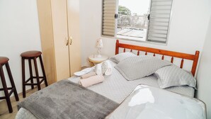 Un dormitorio con una cama, dos taburetes de madera, un armario, una mesita de noche con lámpara y una ventana con persianas.
