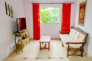 Ein Wohnzimmer mit einer Couch, einem Tisch, einem Fernseher auf einem Schrank und einem Fenster mit roten Vorhängen.