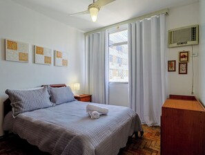 Ein ordentlich eingerichtetes Schlafzimmer mit einem Bett, einem Nachttisch, einem Holznachtkästchen, einem an der Wand befestigten Fernseher und einem Fenster mit Vorhängen.