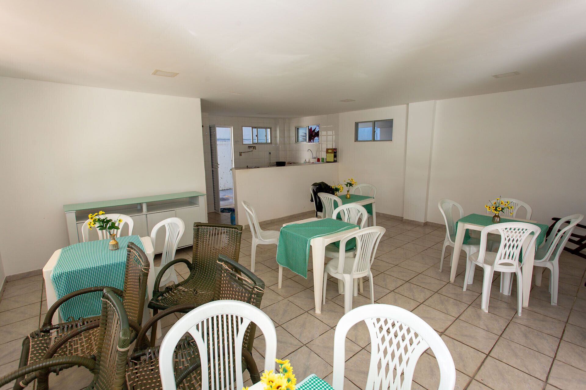 A dining area with white plastic chairs and tables, a wicker chair, and a kitchen area in the background.