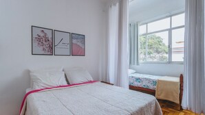 A bedroom with a bed, a small table, and a window with curtains. There are three framed pictures on the wall. - Apto confortável, ótima localização e pet friendly (Teresópolis)