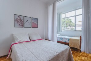 A bedroom with a bed, a small table, and a window with curtains. There are three framed pictures on the wall.