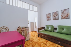 A room with a wooden herringbone floor, a green sofa with a wooden frame, a pink table, wicker chairs, and a white cabinet.