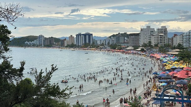 A crowded beach with people swimming and sunbathing, surrounded by tall buildings and a clear sky. - Apto aconchegante c Wi-Fi e varanda para descansar (Caraguatatuba)
