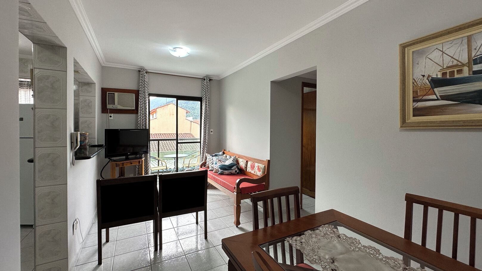 A living room with a TV, a sofa, a dining table, and a balcony with a view of buildings.