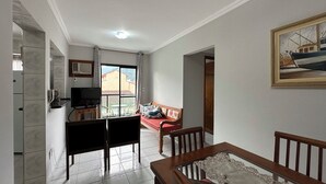 A living room with a TV, a sofa, a dining table, and a balcony with a view of buildings. - Apto aconchegante c Wi-Fi e varanda para descansar (Caraguatatuba)