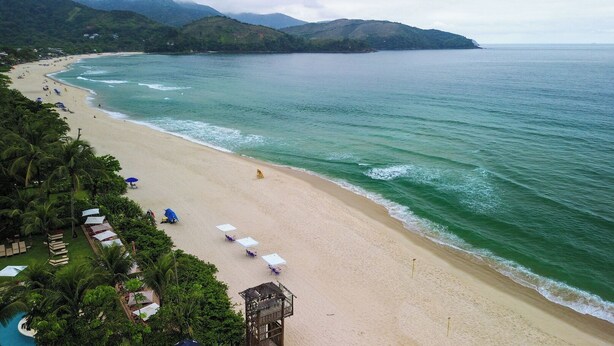 Una playa con una fila de sombrillas blancas y sillas de descanso, una pequeña construcción, y vista al océano y colinas al fondo.