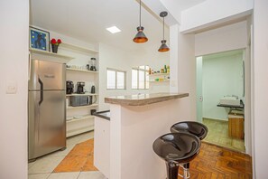 Eine moderne Küche mit integriertem Kühlschrank, Mikrowelle und Kaffeemaschine auf der Arbeitsplatte. Über der Spüle hängen Pendelleuchten und in der Nähe der Arbeitsfläche steht ein kleiner Hocker.