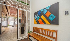 Ein Flur mit einer hölzernen Bank, einem geometrischen Kunstwerk an der Wand und einem weißen Tor mit Pflanzen.