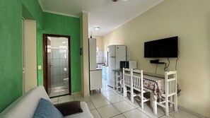 A living room with a sofa, a dining area with a table and chairs, a TV mounted on the wall, and a kitchen area with a refrigerator and microwave. - Apartamento aconchegante c Wi-Fi na Praia do Sapê (Ubatuba)