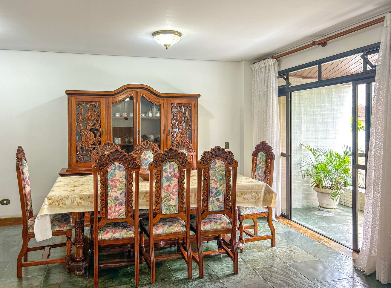 A dining area with a wooden table and chairs, a floral-patterned tablecloth, and a cabinet with glass doors.