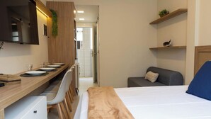 Quarto de hotel com uma cama, uma escrivaninha com cadeira, um pequeno sofá e uma televisão fixada na parede.
