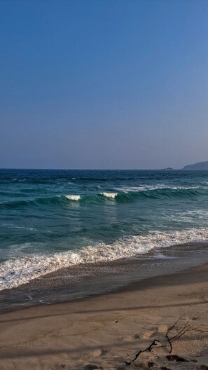 Una playa con olas acercándose a la orilla, cielo despejado y formaciones terrestres a lo lejos.