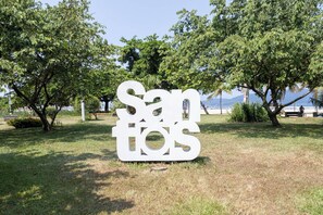 Um grande letreiro branco com o texto "Sanitos" em um parque com árvores e uma praia ao fundo.