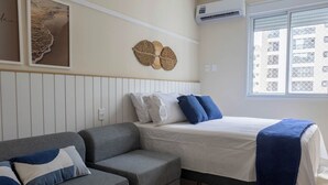 A bedroom with a bed, a gray sofa, a wall-mounted air conditioner, and a window with a view of buildings. - Apartamento Moderno com Wi-Fi em frente a praia (Santos)