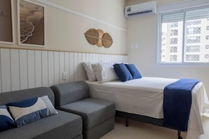 Quarto com cama, sofá cinza, ar-condicionado fixado na parede e janela com vista para edifícios.
