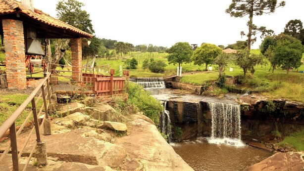 A rustic stone bridge leading to a wooden structure with a tiled roof, overlooking a small waterfall and a lush green landscape.