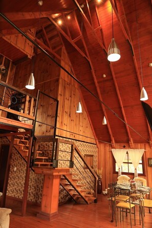 Une pièce spacieuse avec un plafond haut, des poutres en bois, un escalier et un coin repas comprenant une table et des chaises.