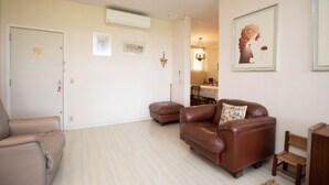 Een woonkamer met een bruine leren bank, een beige fauteuil en een kleine houten tafel. Er hangen ingelijste foto's aan de muur en er is een kroonluchter op de achtergrond.