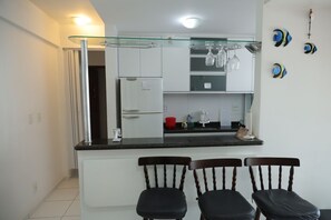 A compact kitchen with a refrigerator, a glass shelf with wine glasses, and a dining area with two chairs. - Ótimo Apto à 650m da praia do Forte em Cabo Frio (Cabo Frio)