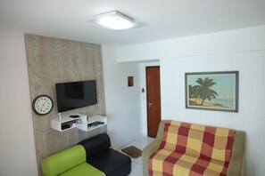 A living room with a flat-screen TV, a clock, a shelf with items, a green and black sofa, and a colorful armchair. - Ótimo Apto à 650m da praia do Forte em Cabo Frio (Cabo Frio)